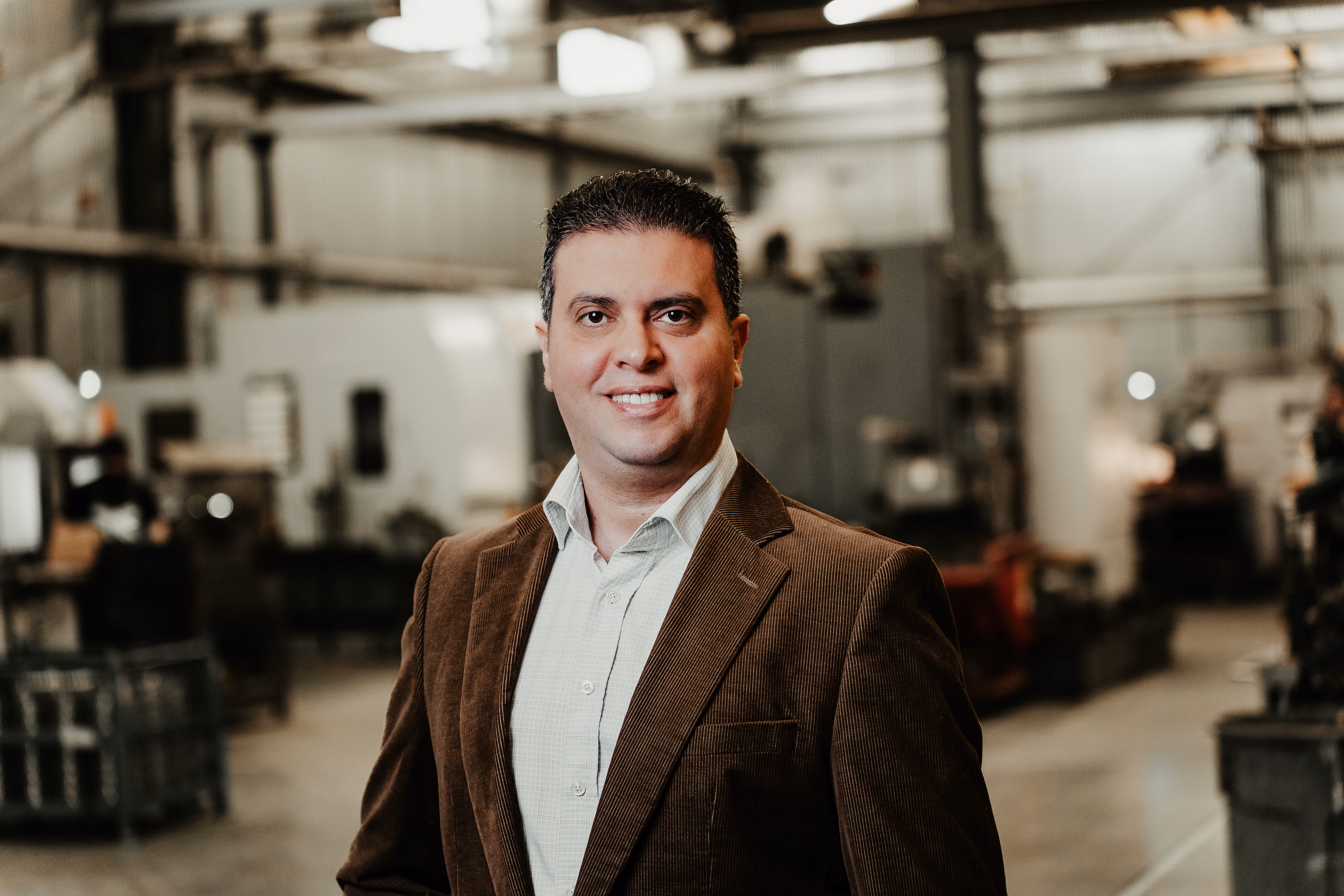 Man in a brown blazer and white shirt smiling, standing inside an industrial or workshop setting with blurred machinery in the background.