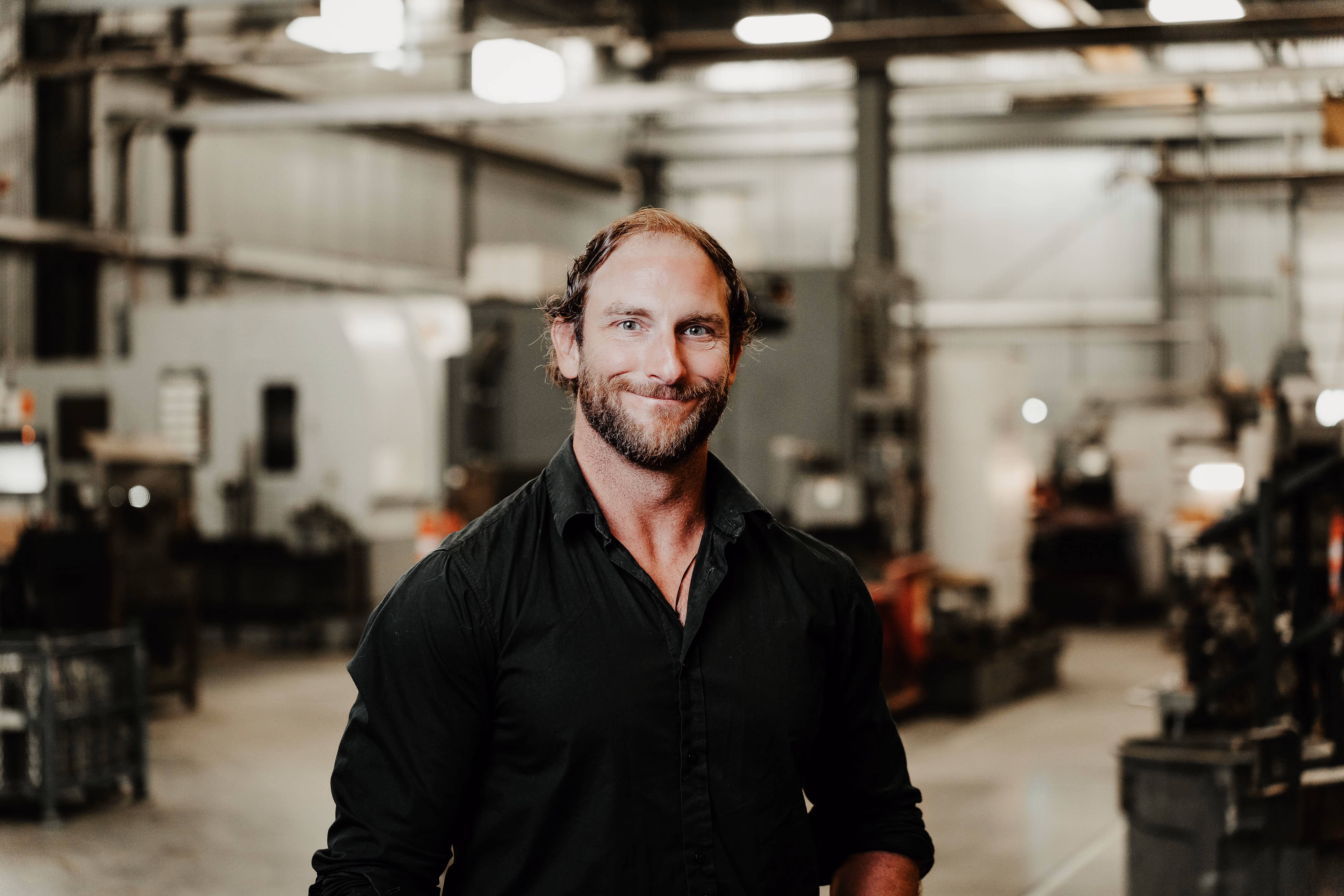A man with a beard and short hair, wearing a black button-up shirt, standing and smiling inside a spacious industrial warehouse with machinery and equipment in the background.