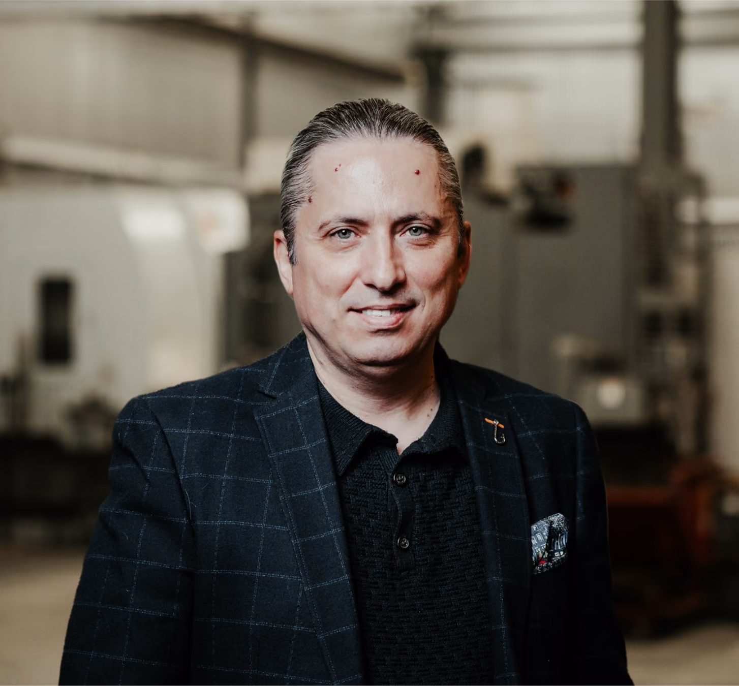 Man wearing a black checkered blazer and a black shirt stands smiling in an industrial indoor setting with blurred machinery in the background.
