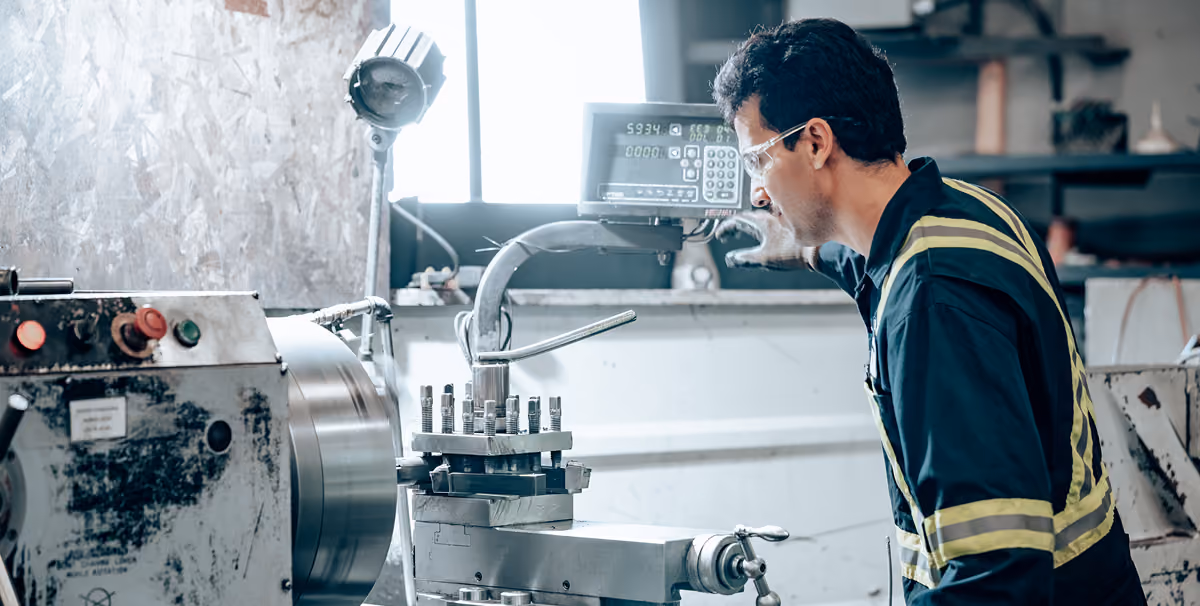 Man wearing safety glasses and a striped work shirt operating a metal lathe machine in an industrial workshop.