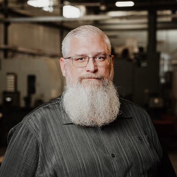 Portrait of a man with glasses and a long white beard wearing a dark button-up shirt in an industrial or workshop setting.
