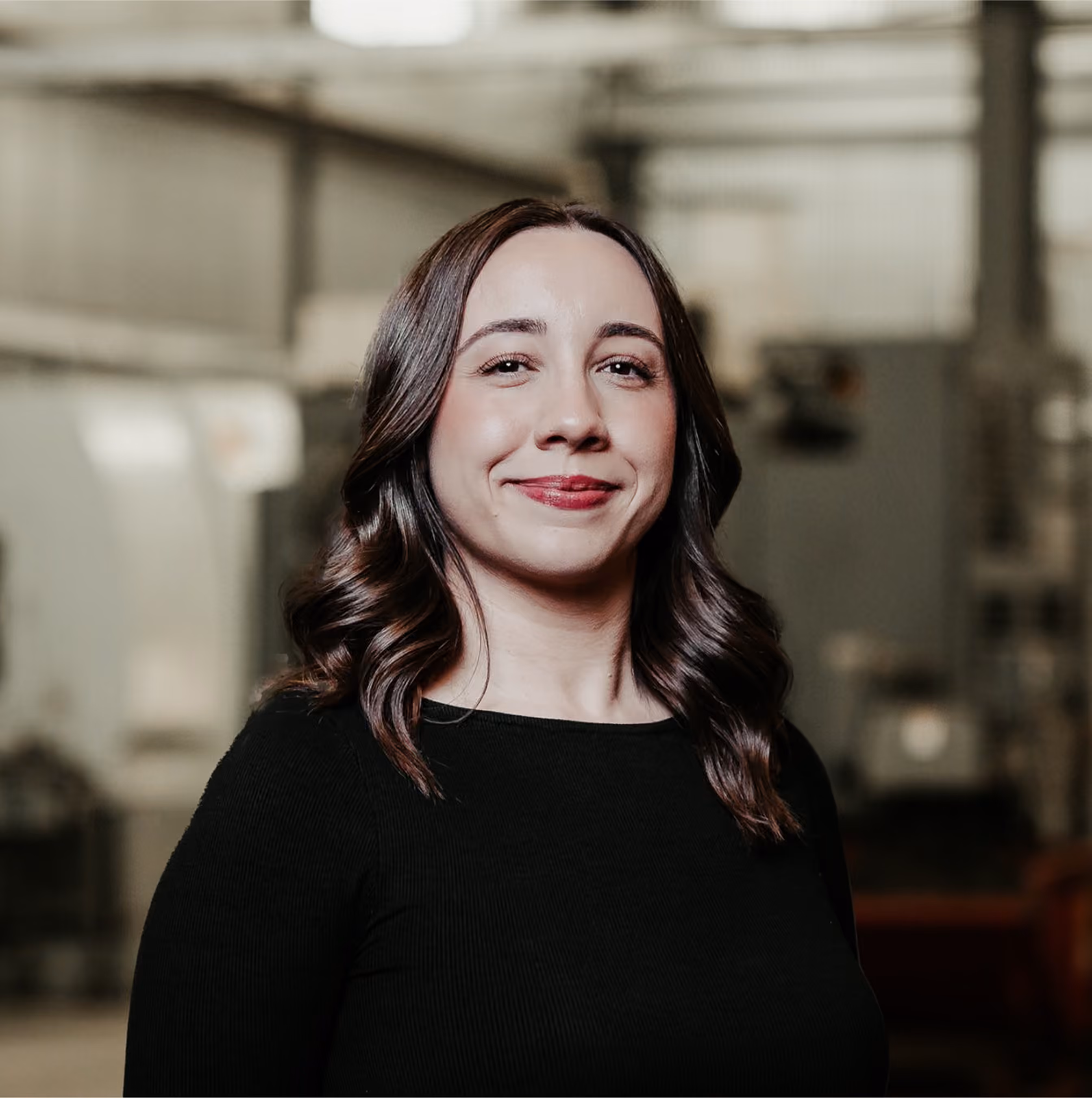 Smiling woman with wavy dark brown hair wearing a black top, standing indoors with industrial background.