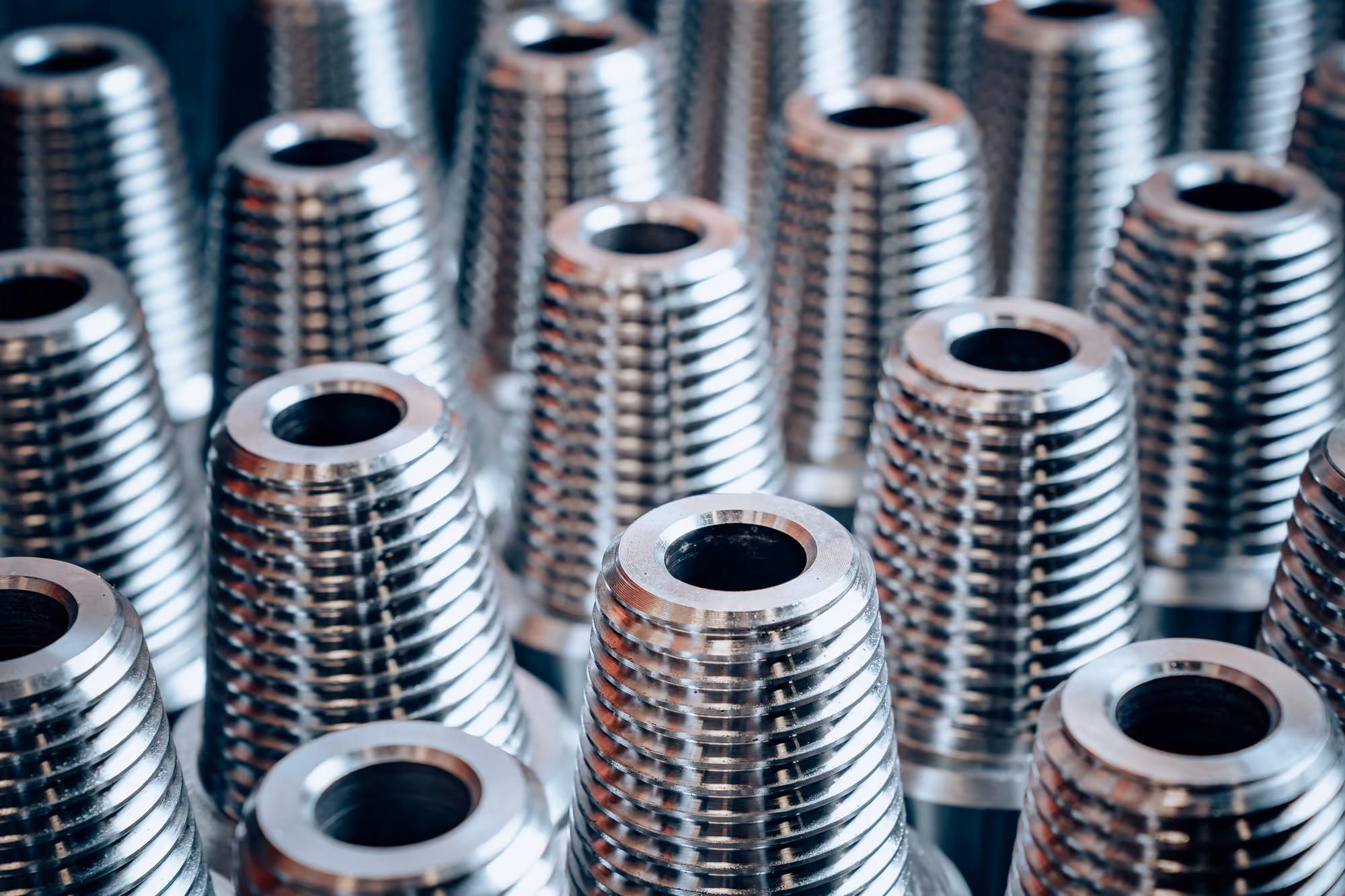 Close-up of multiple shiny metal threaded inserts arranged in rows, showing their cylindrical ridged surfaces and hollow centers.