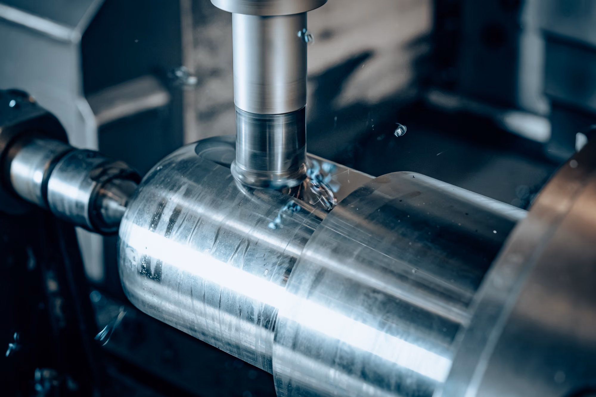 Close-up of a CNC milling machine cutting a cylindrical metal workpiece, with metal shavings visible around the cutting tool.
