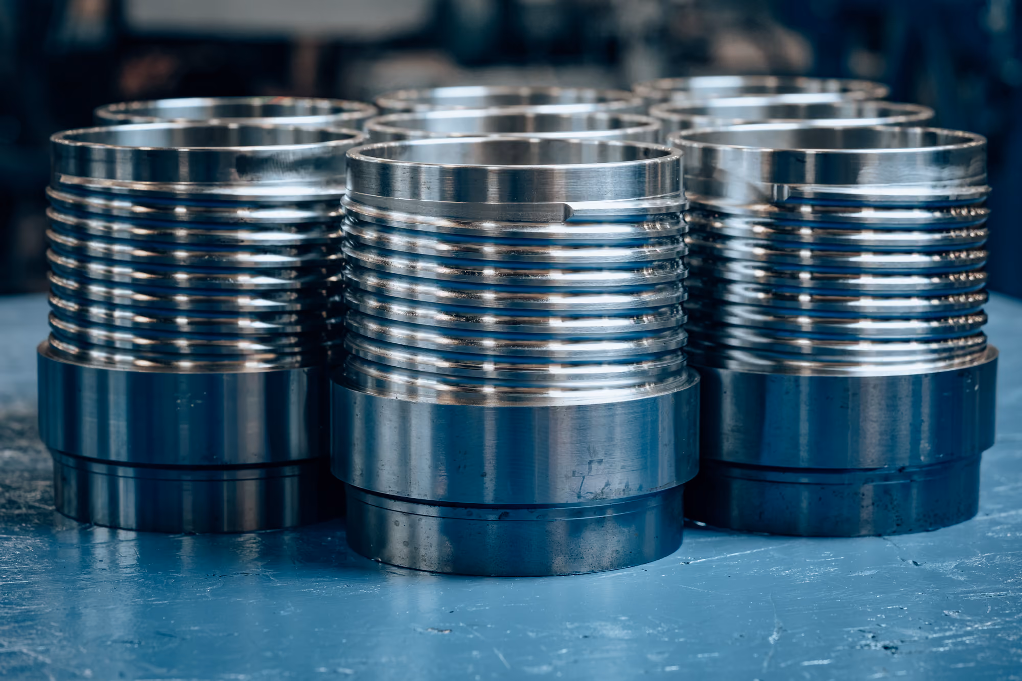 Close-up of several large, cylindrical metal components with precision threading, arranged in rows on a blue surface.