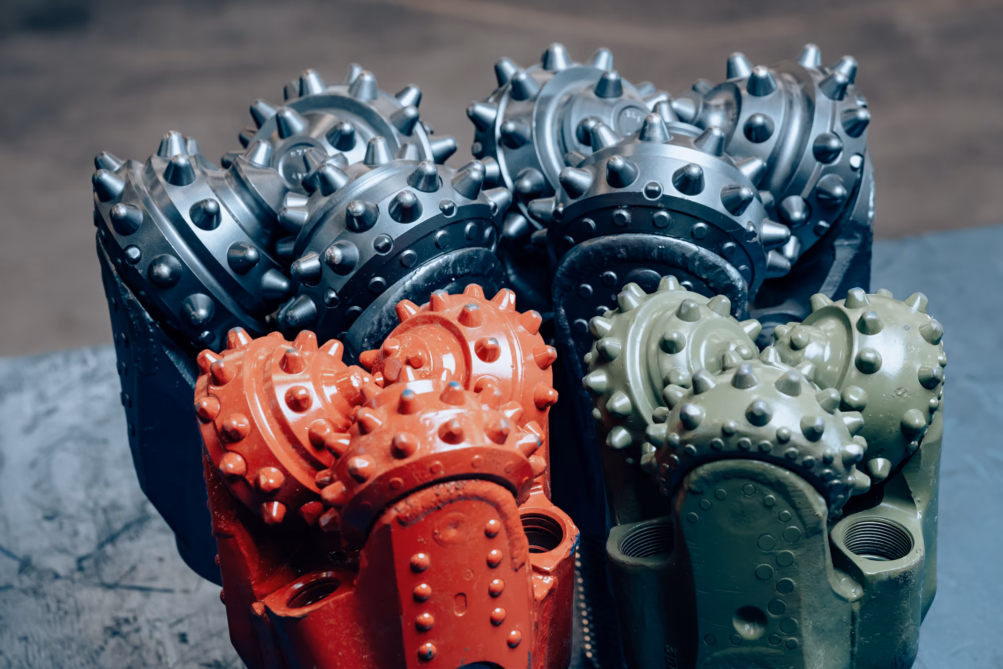 Close-up of three tricone drill bits in black, red, and green resting on a surface, used for drilling in construction or mining.