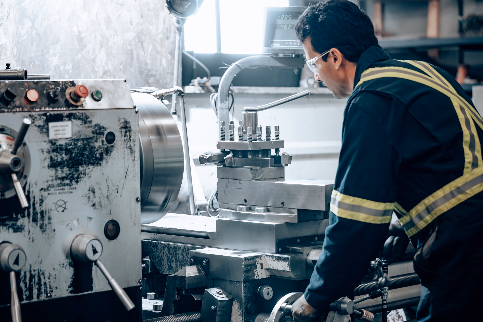 Man wearing safety glasses and a dark uniform with reflective stripes operating a metal lathe machine in an industrial workshop.