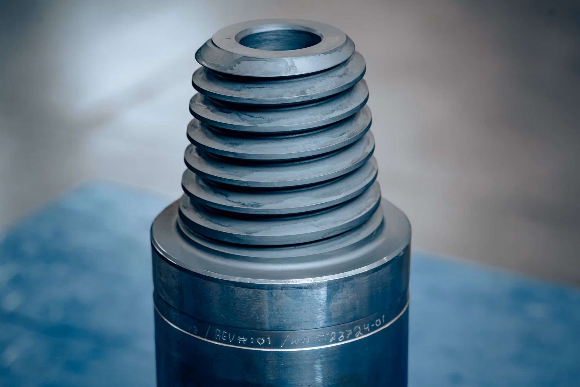 Close-up of a large metal screw with a round hollow top and thick helical threads, set on a blue surface with a blurred background.