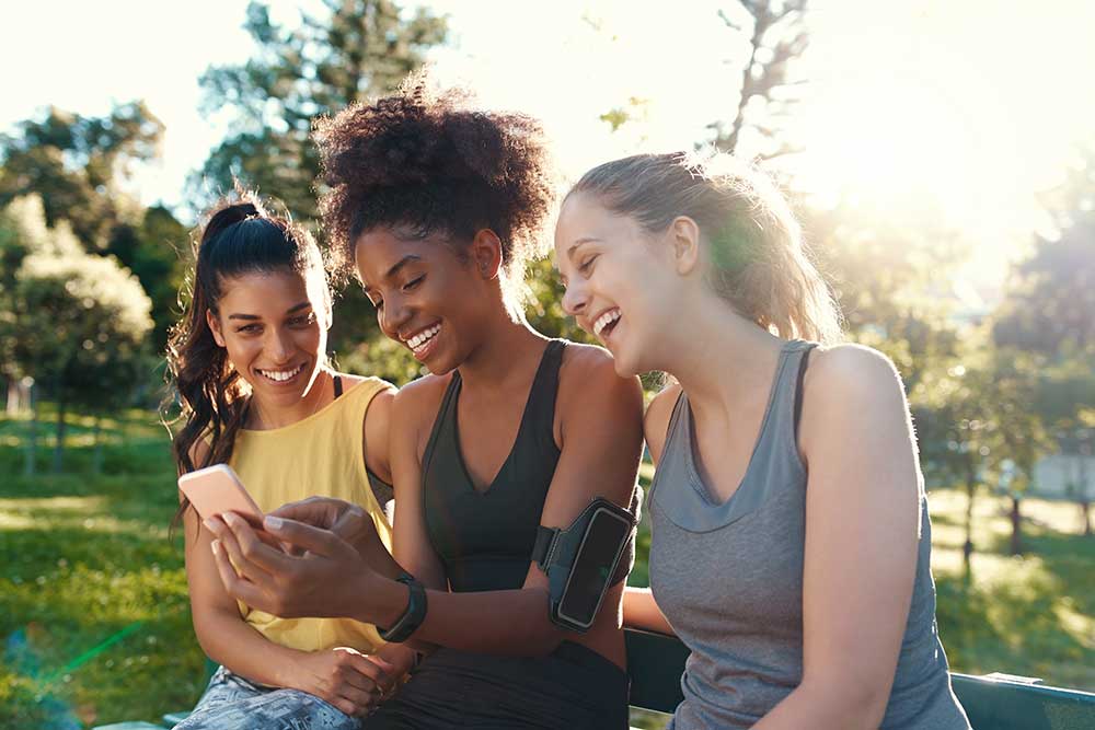Imagem de 3 mulheres se exercitando ao ar livre e olhando um smartphone.