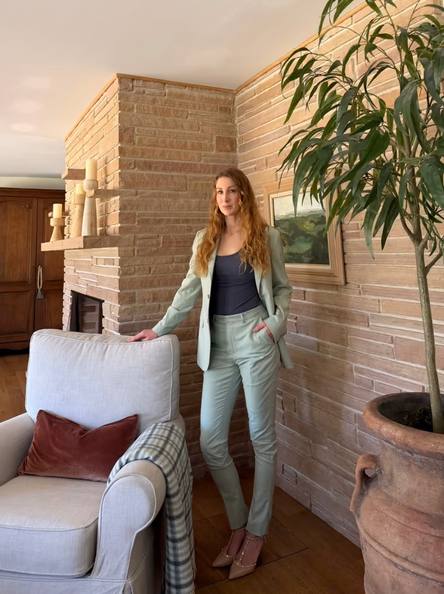 Woman in light green suit with black top standing near a sofa and brick fireplace in a cozy room.