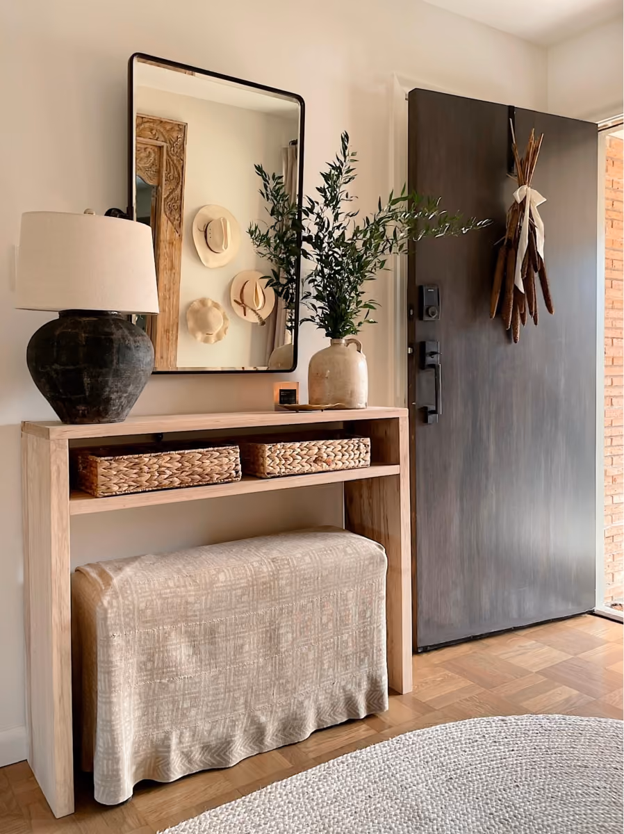 Entryway with wooden console table, lamp, green plant in vase, woven baskets, mirror, and dark front door wreath.