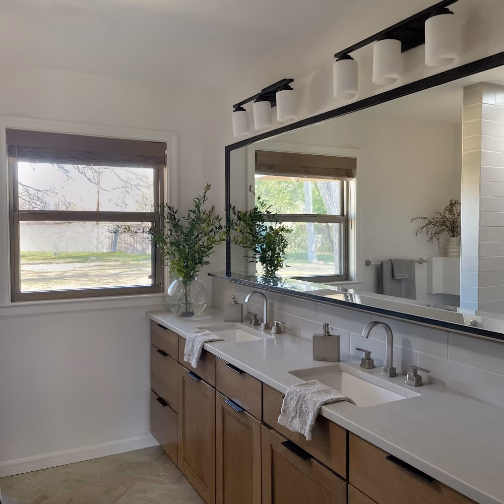 Contemporary bathroom with dual sinks, large mirror, wooden cabinets, and a window with a view outside.
