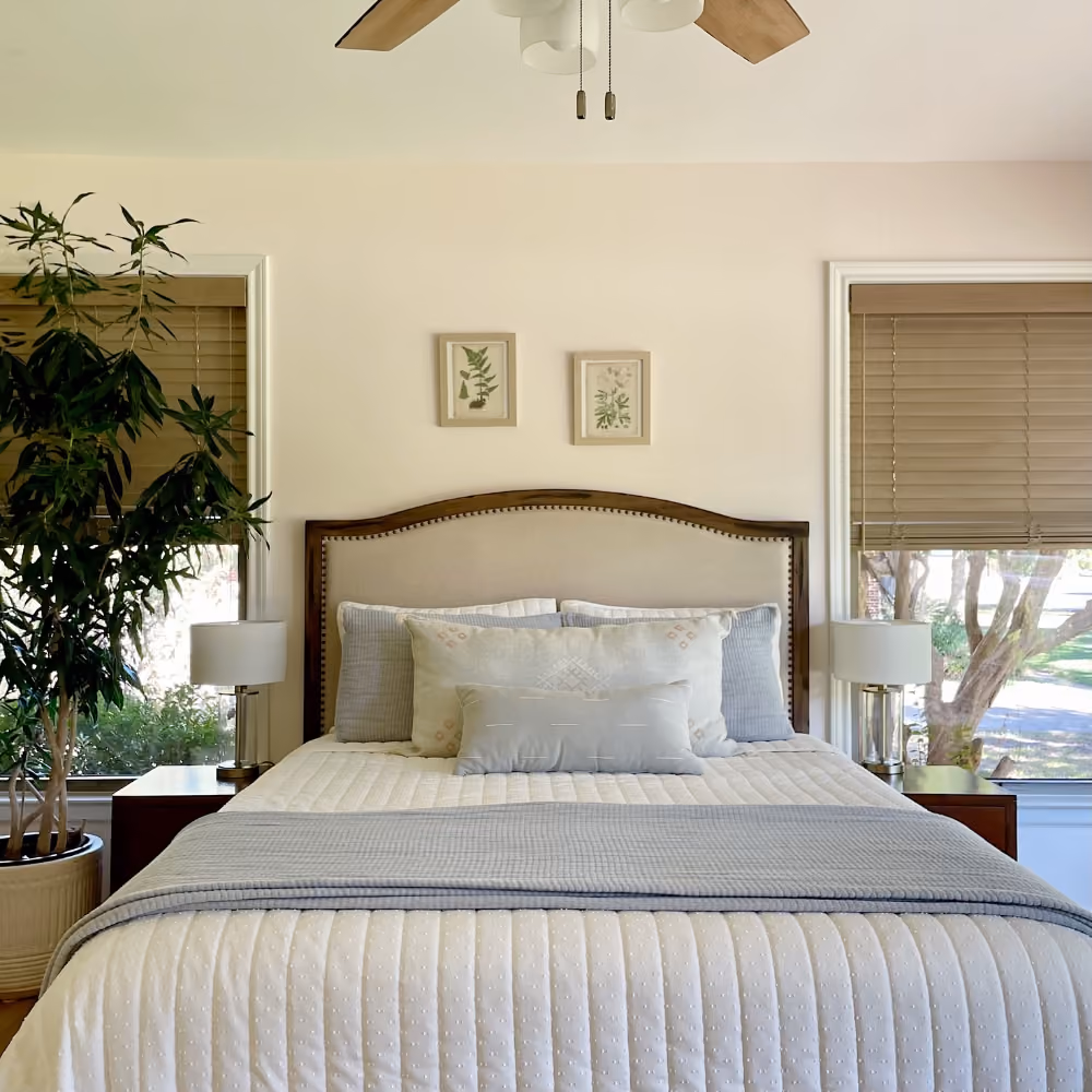 Cozy bedroom with a bed, light bedding, two pillows, wooden headboard, nightstands, lamps, and window blinds.