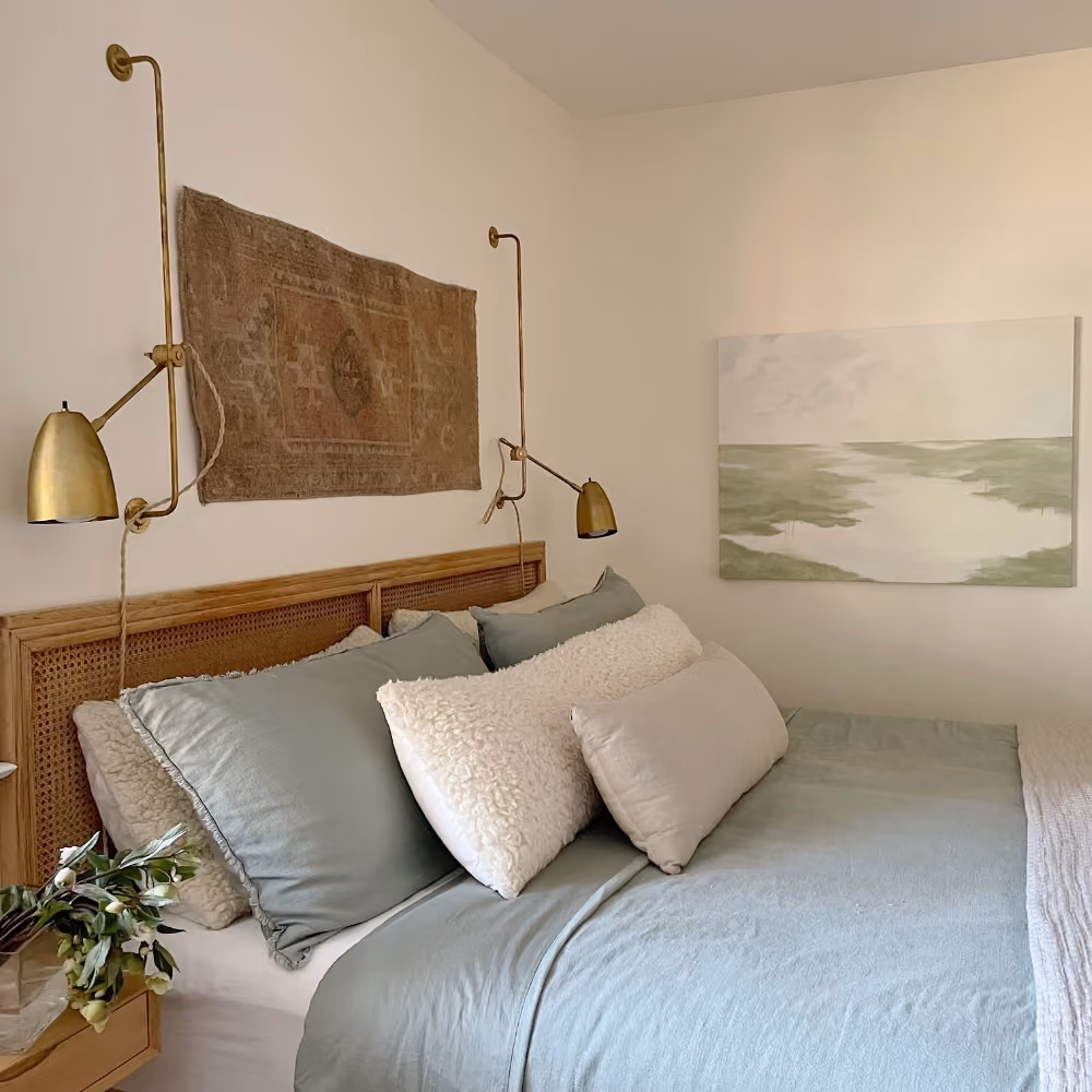 Calm bedroom with light blue bedding, textured pillows, brass lamps, and art on the cream walls.