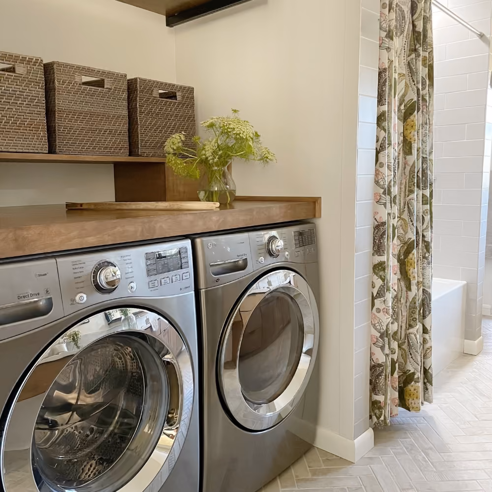 Laundry room with silver washer, dryer under wooden countertop and storage baskets on shelf above.