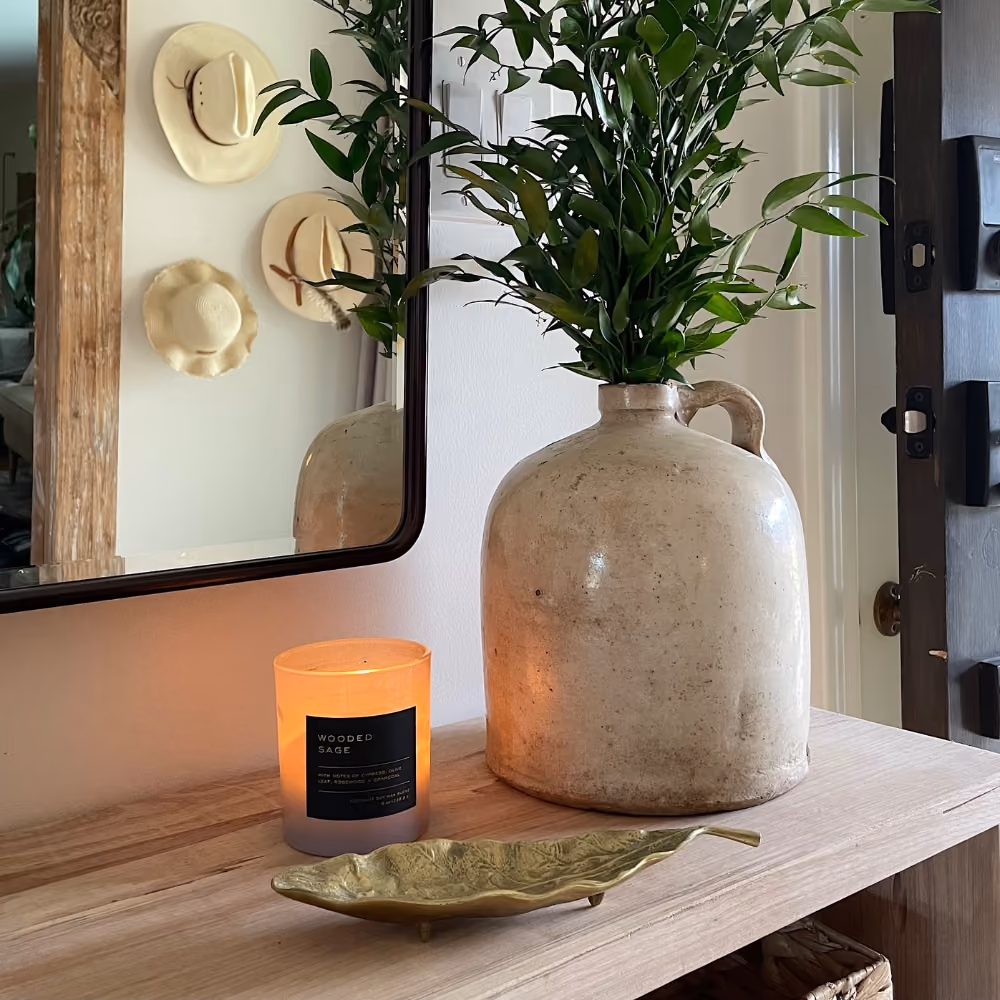 Wooden entry table with a lit Wooded Sage candle, ceramic jug with green leaves, and brass leaf tray under a mirror.