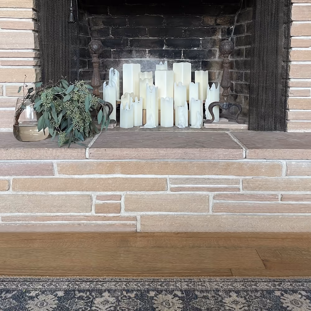 Fireplace filled with unlit white candles and rustic metal andirons, with greenery in a glass vase.