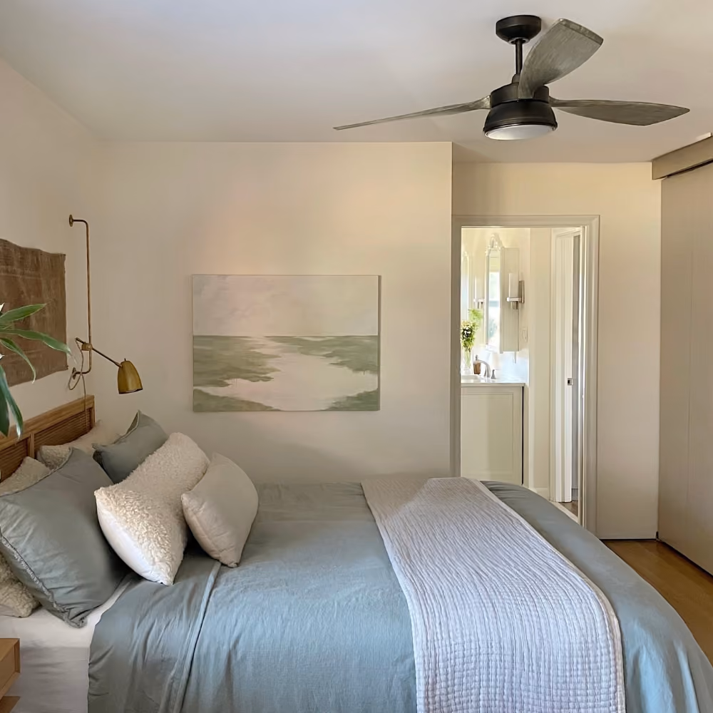 Cozy bedroom with blue bedding, white pillows, a ceiling fan, wall lamp, and abstract landscape painting.