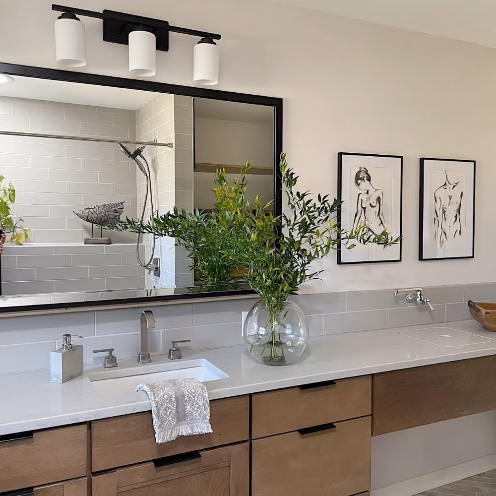 Modern bathroom with wooden vanity, white countertop, large mirror, green plant, and black framed art.
