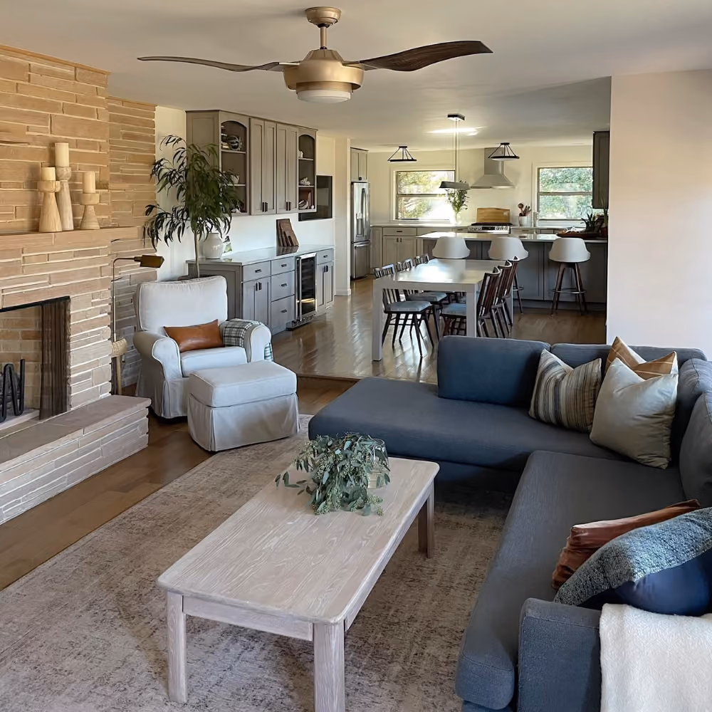 Open living room with gray sectional sofa, armchair, wooden coffee table, brick fireplace, and kitchen in background.