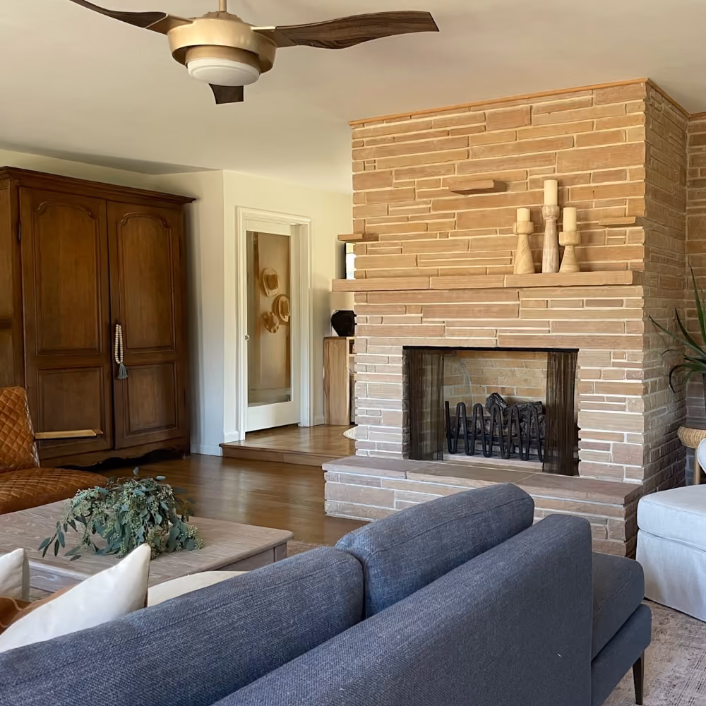 Cozy living room with a stone fireplace, gray sofa, wooden cabinet, ceiling fan, and coffee table with greenery.