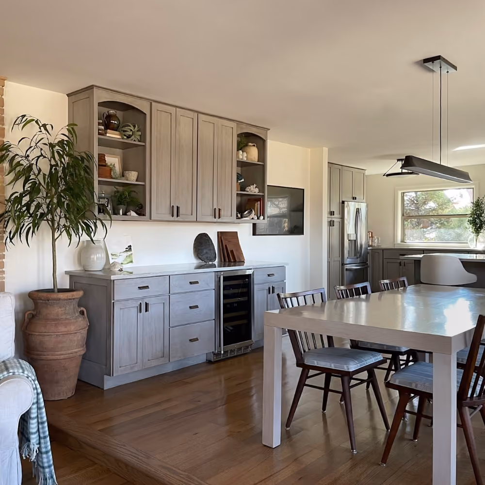 Modern dining room with wooden table, chairs, gray cabinetry, potted plant, and stainless steel refrigerator.