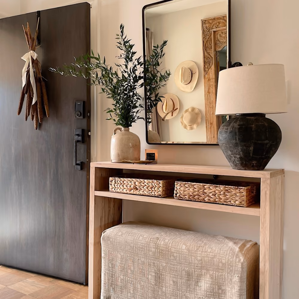 Wooden entryway table with a lamp, plant in vase, two baskets, and mirror reflecting wall hats.