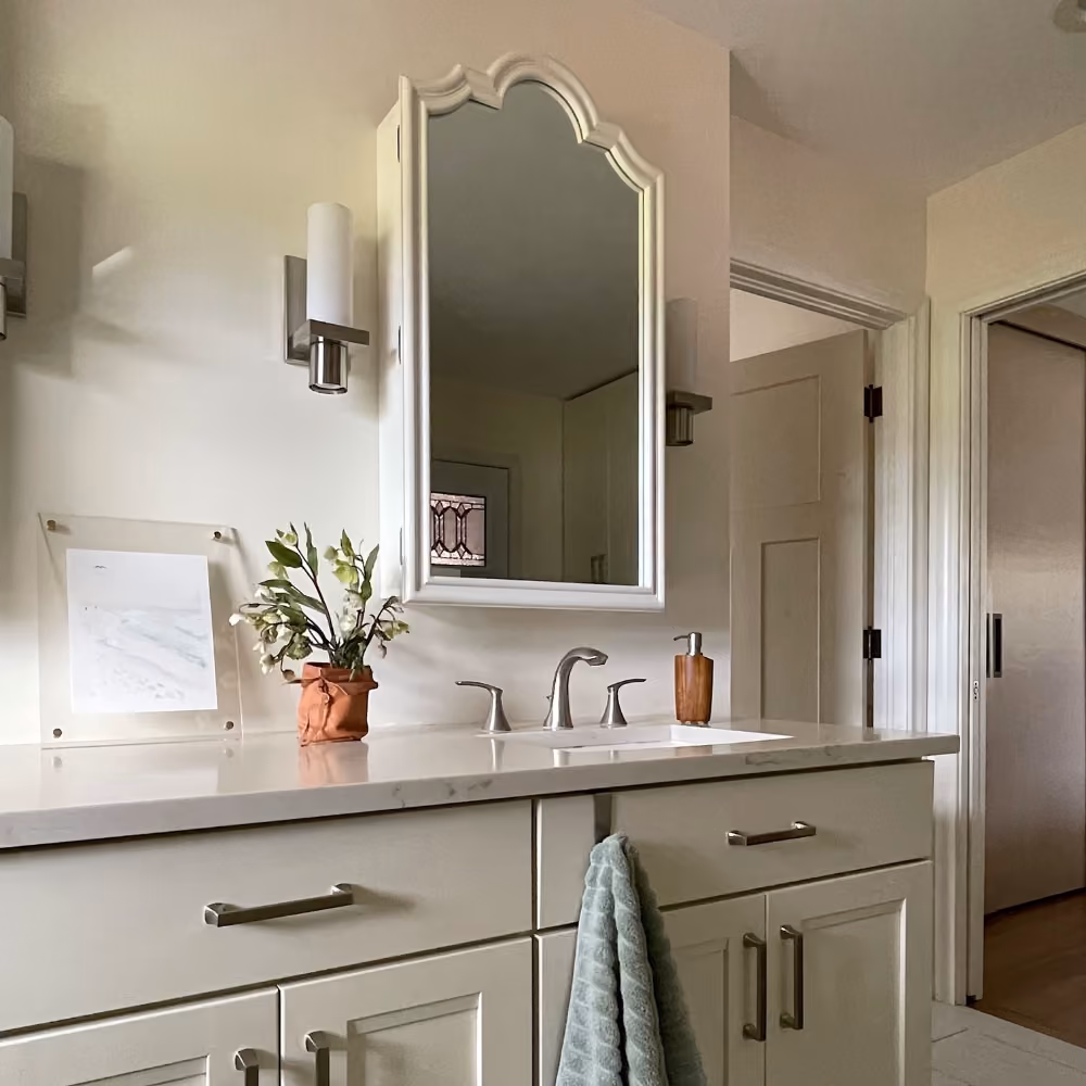 Bathroom vanity with a white counter, floral arrangement, soap dispenser, and arched mirror above sink.