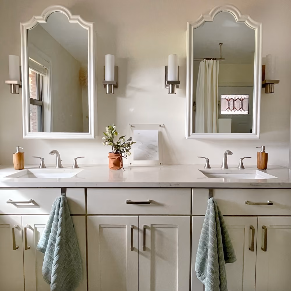 Double white bathroom vanity with two mirrors, sinks, silver faucets, and green towels hanging on cabinet handles.
