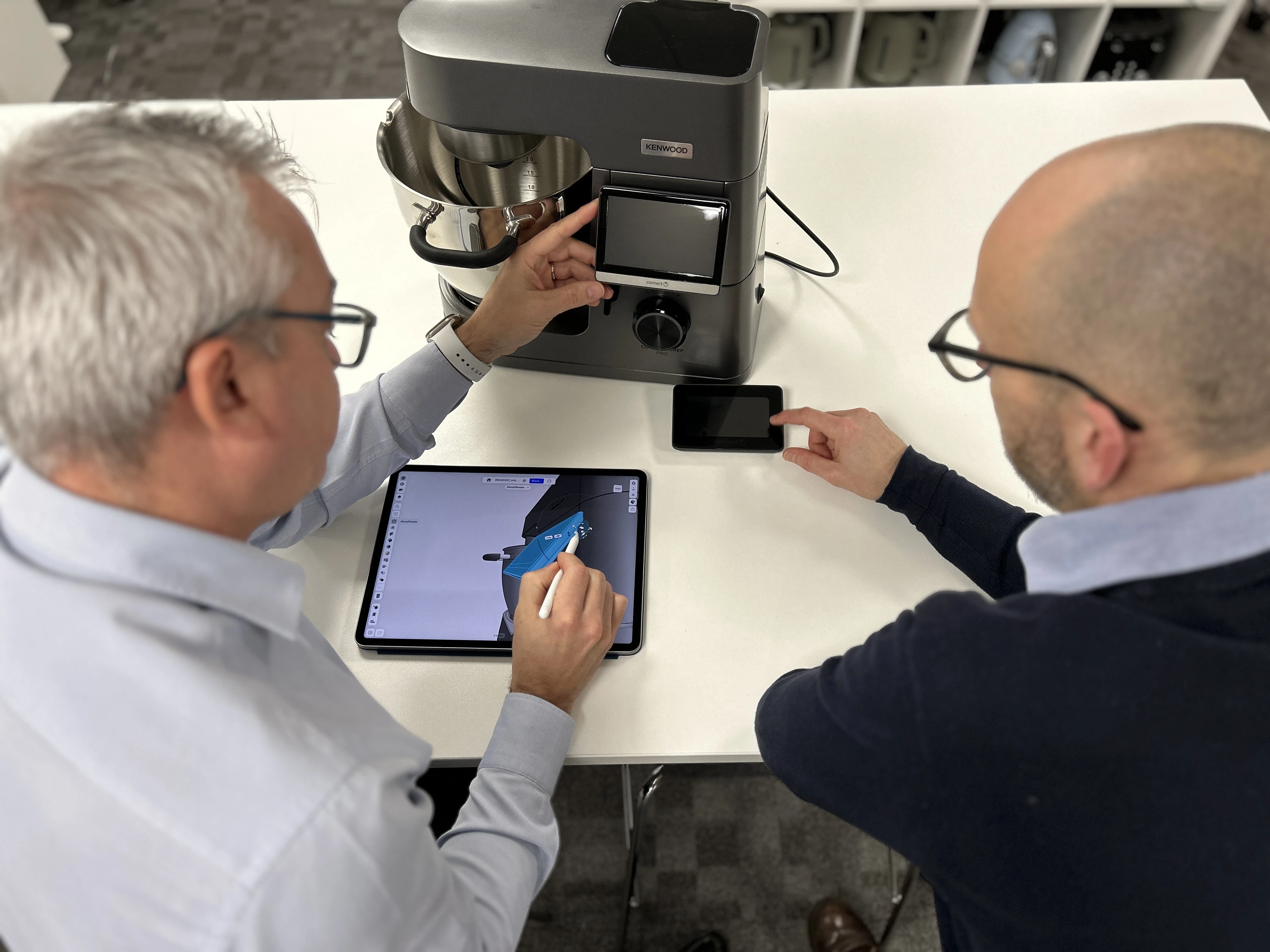 Two designers sit at a desk with a standing mixer appliance in front of them. They are modeling a part of the appliance in Shapr3D on an iPad with the Apple Pencil.