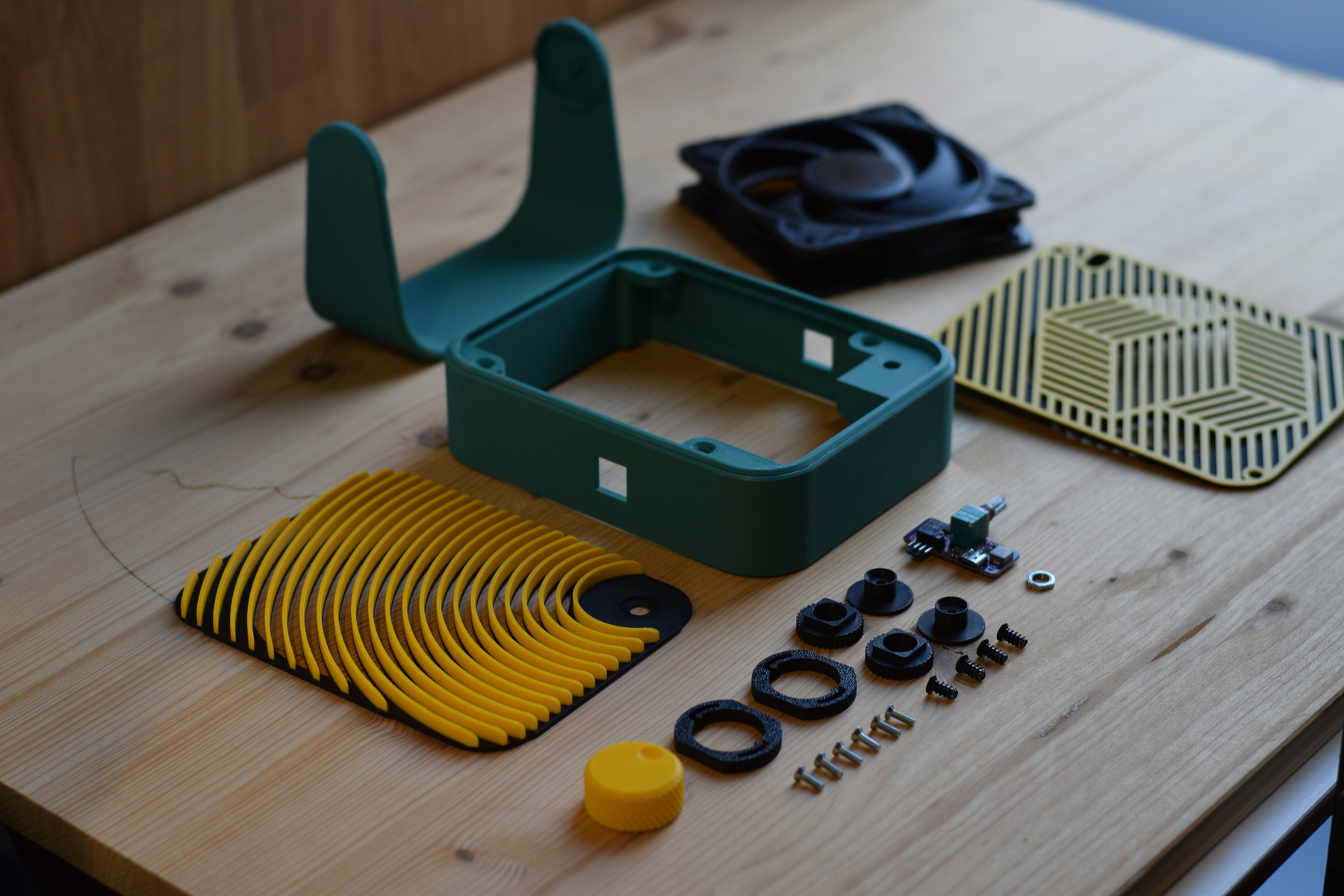 All parts of a 3D printed USB-C desk fan laid out: teal body and leg, yellow front grille, back cover, 120 mm PC fan, USB-C PWM controller board, hinge cylinders and springs, knob, and M3 screws