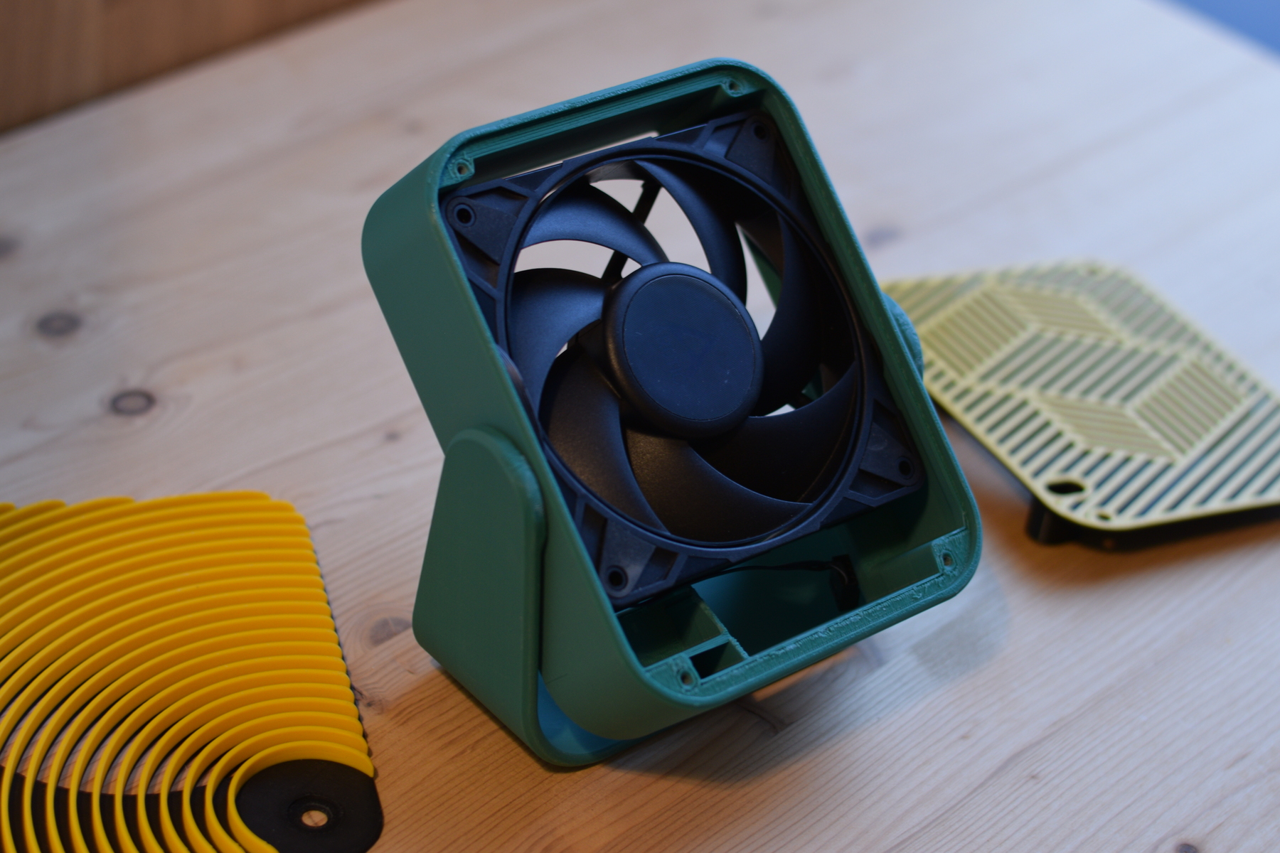 A partially assembled 3D printed desk fan on a wooden table, featuring a black PC fan mounted inside a teal 3D printed housing and leg stand, with yellow and black patterned front and rear grilles lying beside it.