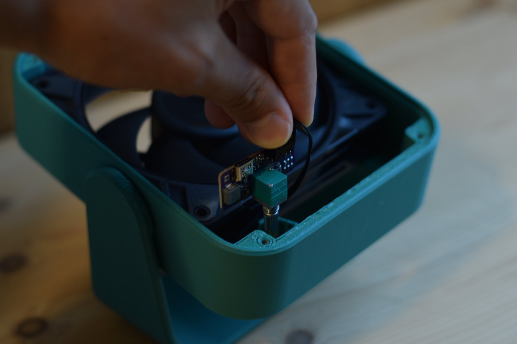 A close-up of a person's hand installing a small USB-C PWM controller board into the teal housing of a partially assembled 3D printed desk fan.