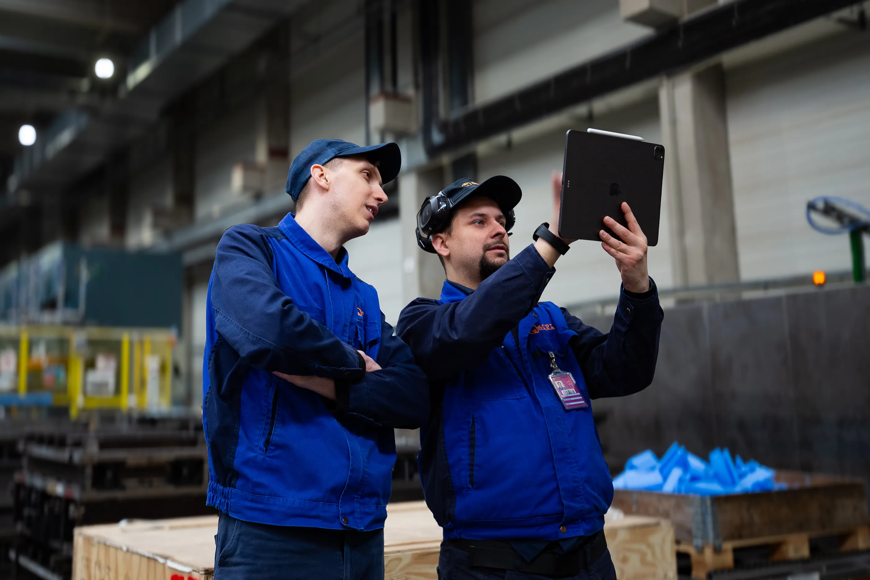 Two workers in blue uniforms and caps stand in an industrial warehouse, with one holding up a tablet running Shapr3D, they both look at the screen.