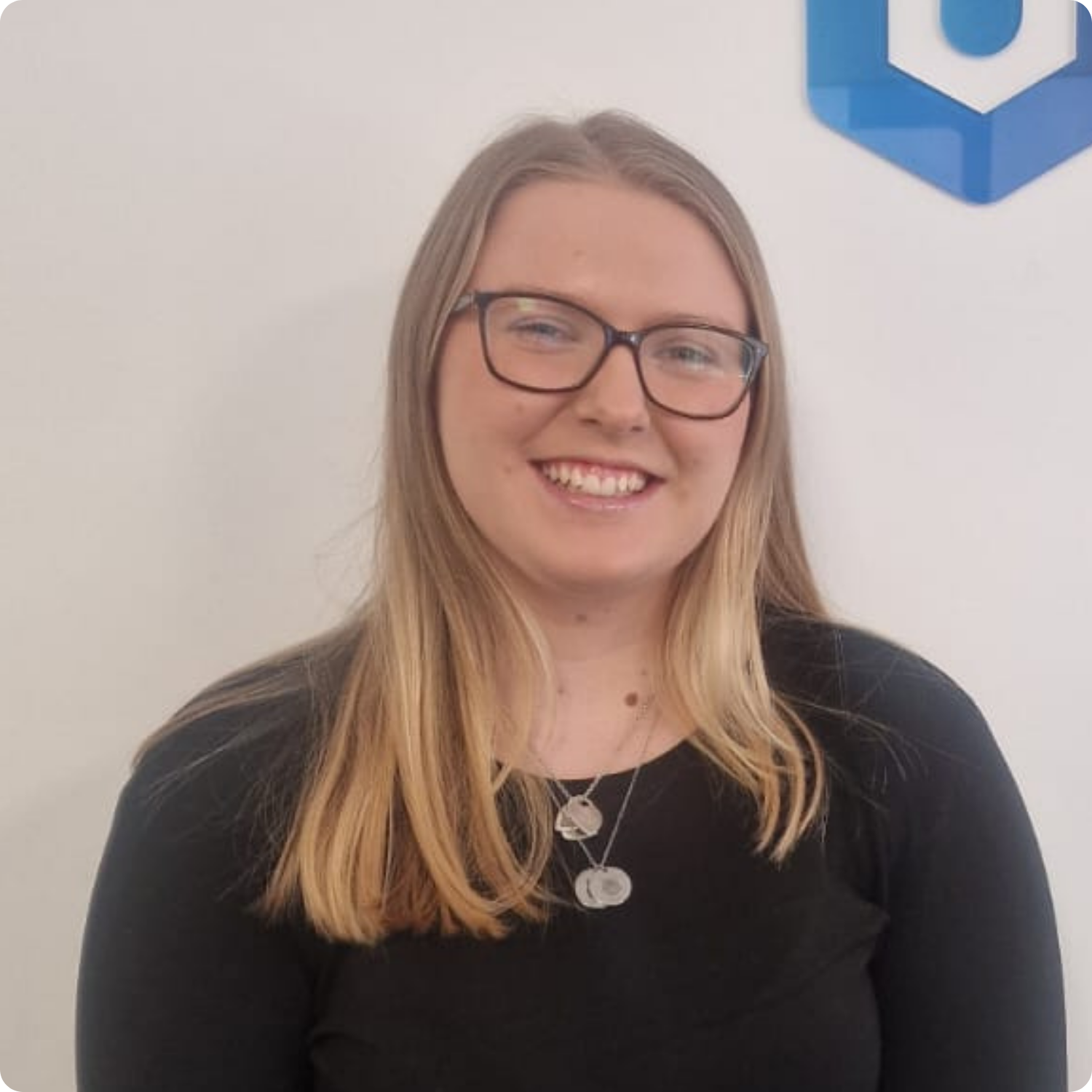 A person with glasses and long hair smiles while standing indoors.