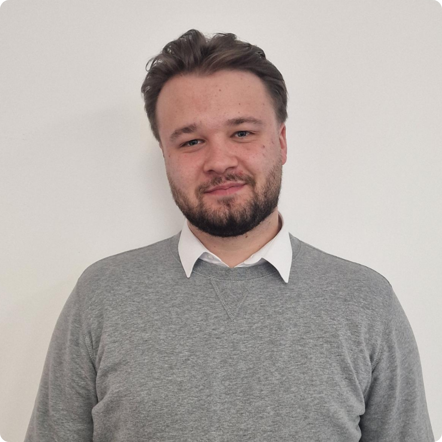 A person in a grey sweater and white shirt stands smiling against a plain white background.