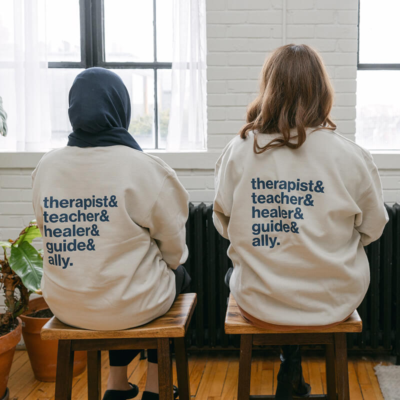 Two women sitting faced away from the camera weaing a sweatshirt that says "therapists & teachers & healers & guides & ally"