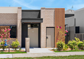Modern brick house entrance with black door, wooden slat feature, and landscaped garden with pink flowering plants.