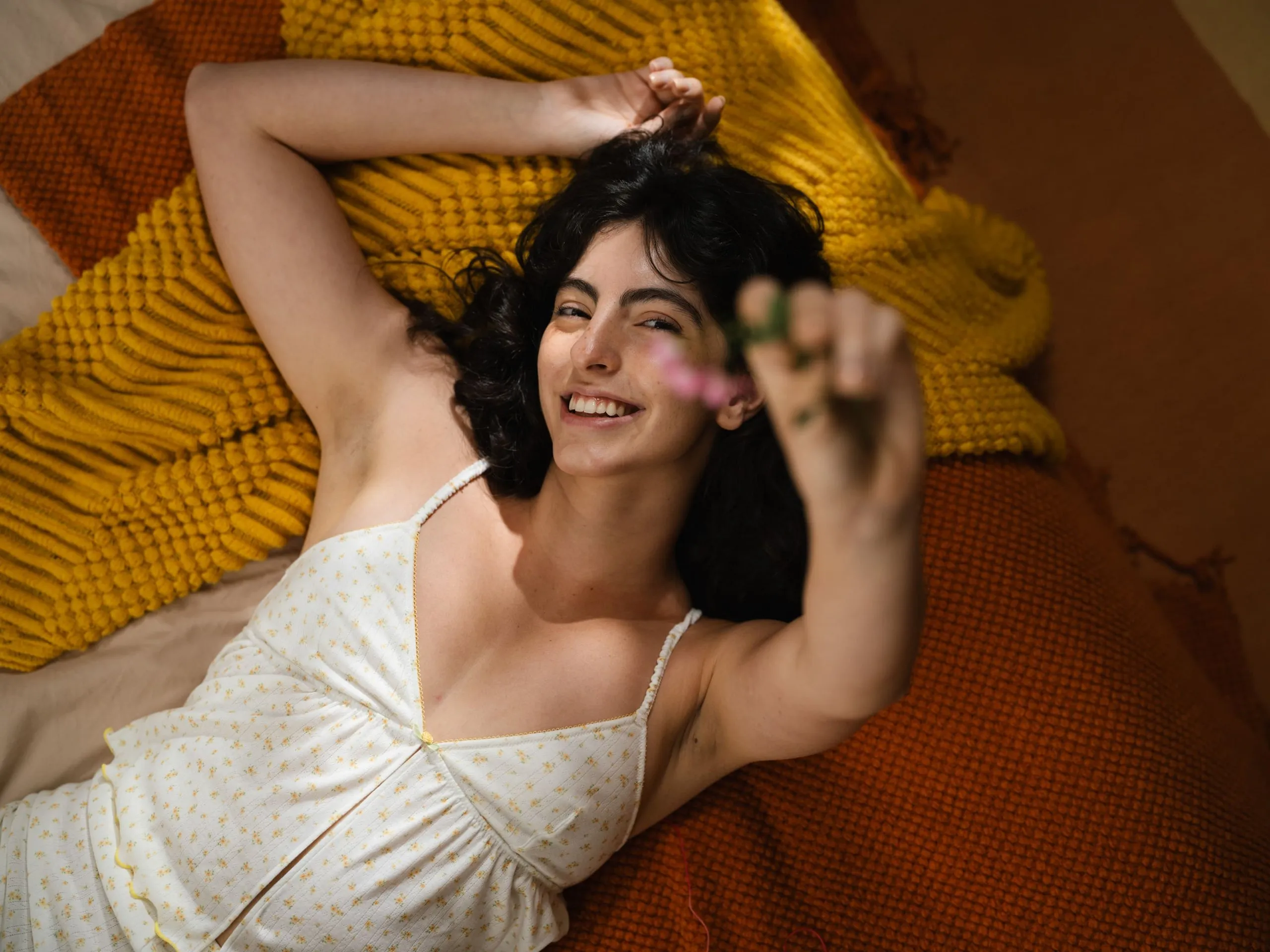 Smiling woman lying on textured orange and yellow blankets reaching forward with a blurred pink flower in hand.