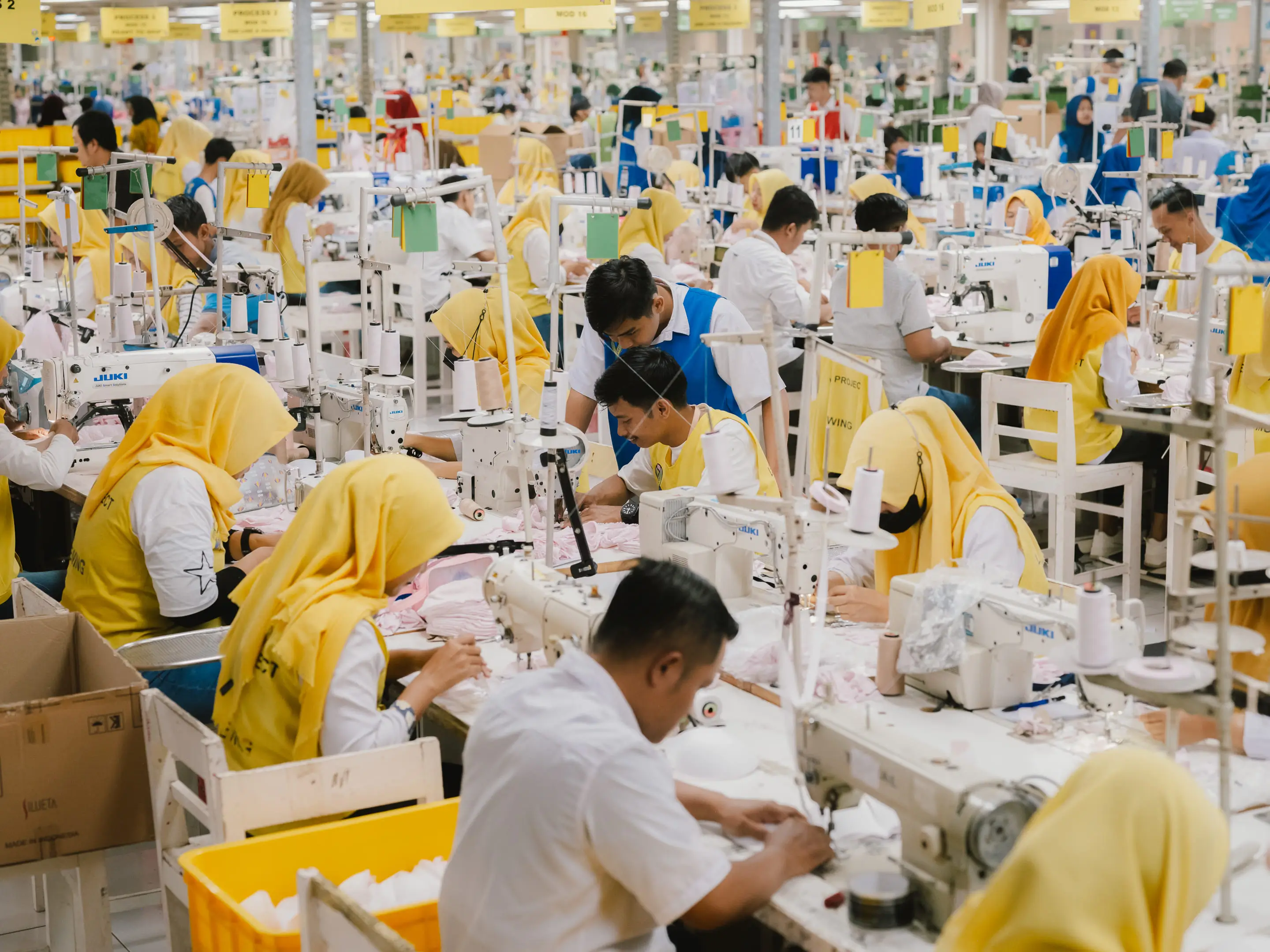 Tailoring factory with workers wearing yellow headscarves and white shirts operating sewing machines.