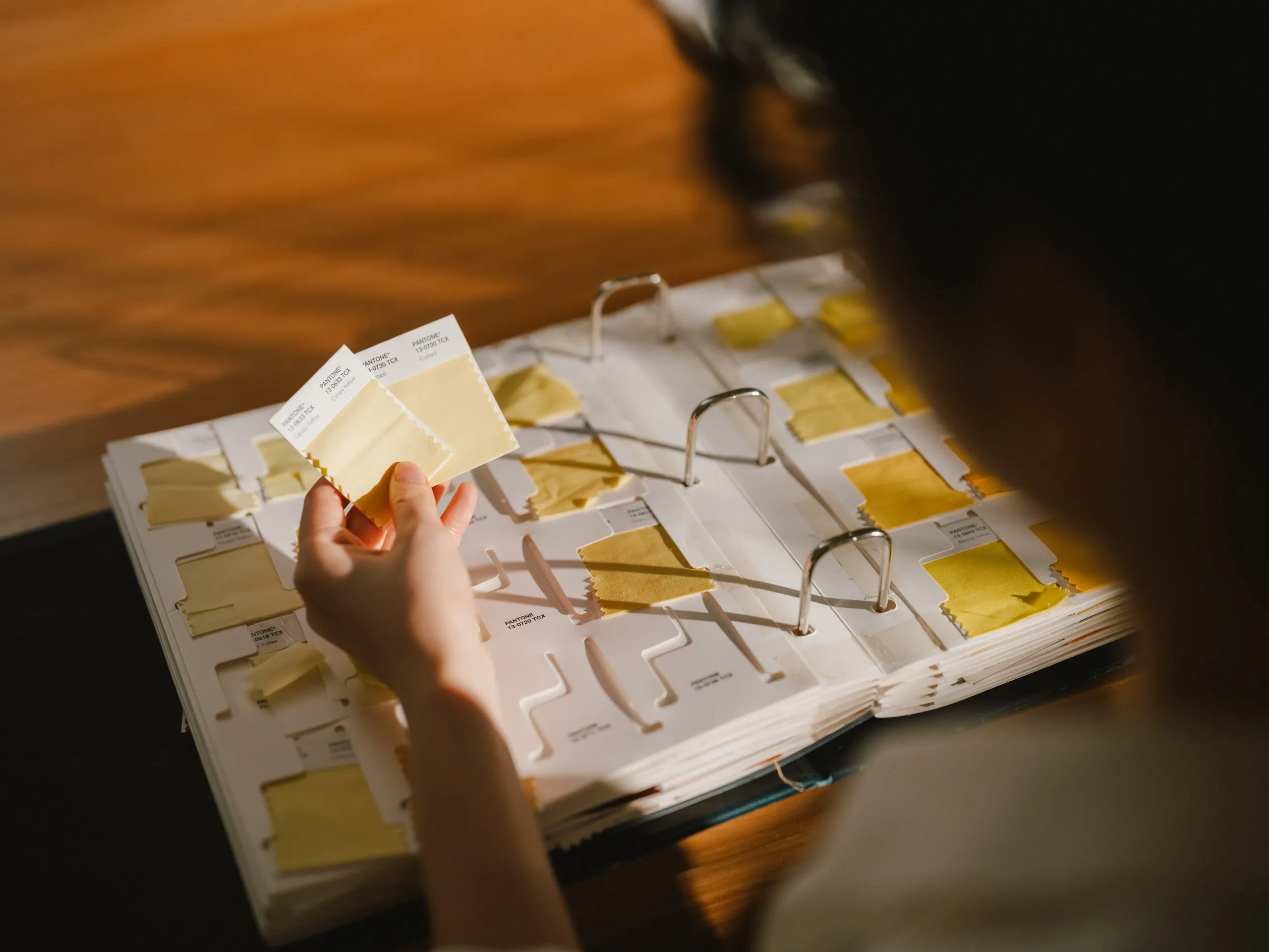 Person holding three Pantone color swatches in shades of yellow over a binder with multiple yellow fabric samples.