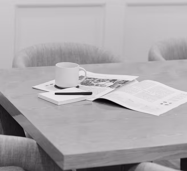 Black and white photos of a meeting space with a coffee mug, newspaper, and pen on a wooden table, with upholstered chairs visible in soft focus background.