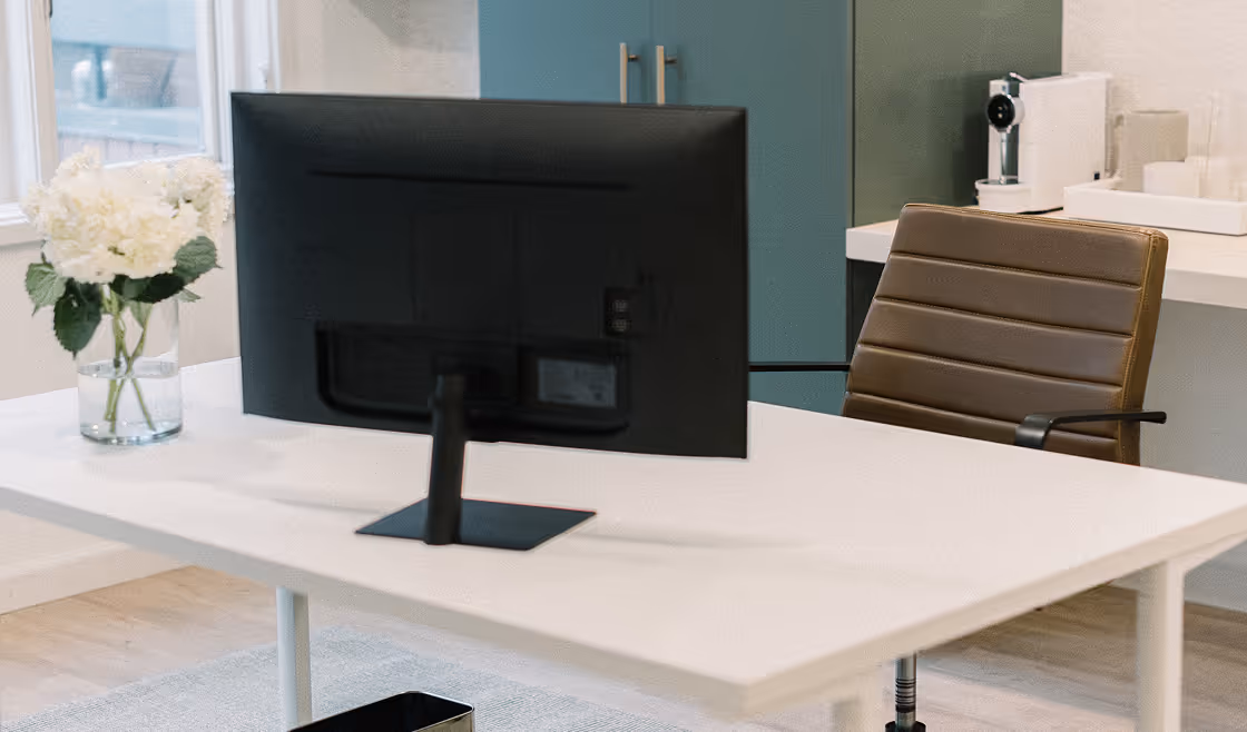 Modern office space with white desk, brown leather office chair, black computer monitor, white flowers in vase, metallic waste basket on floor, and teal cabinet storage in background.
