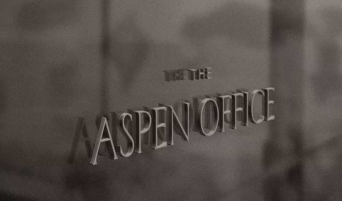 Close-up of raised metallic lettering reading "THE ASPEN OFFICE" mounted on a textured wall surface, photographed in black and white.