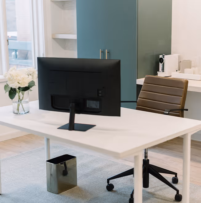 Modern office space with white desk, brown leather office chair, black computer monitor, white flowers in vase, metallic waste basket on floor, and teal cabinet storage in background.