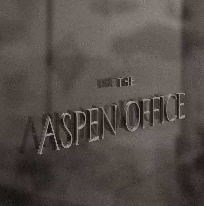 Close-up of raised metallic lettering reading "THE ASPEN OFFICE" mounted on a textured wall surface, photographed in black and white.