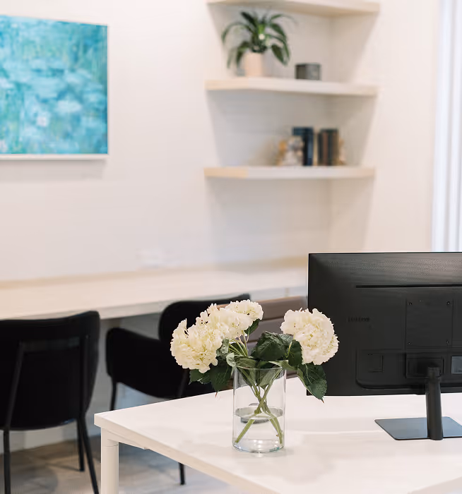 Modern office space with white desk, black computer monitor, white flowers in vase, and additional office chairs in background.