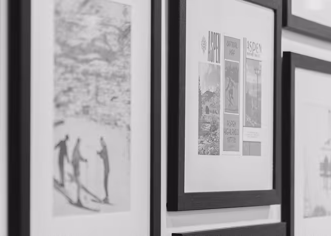 Close-up black and white view of gallery wall showing multiple framed Aspen-inspired pieces in dark frames.