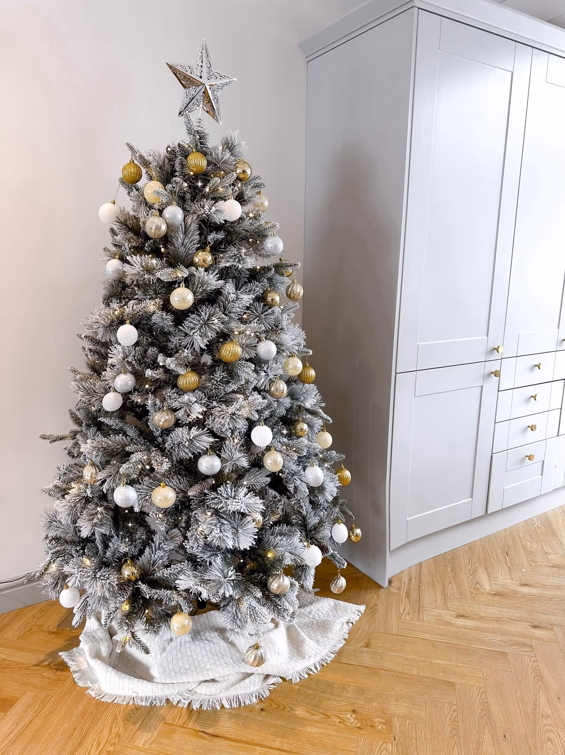 A beautifully decorated 6 ft pre-lit snow flocked mixed pine Christmas tree in a cozy kitchen/dining room. The tree features warm white lights, snowy branches, and pinecones for a realistic winter look.