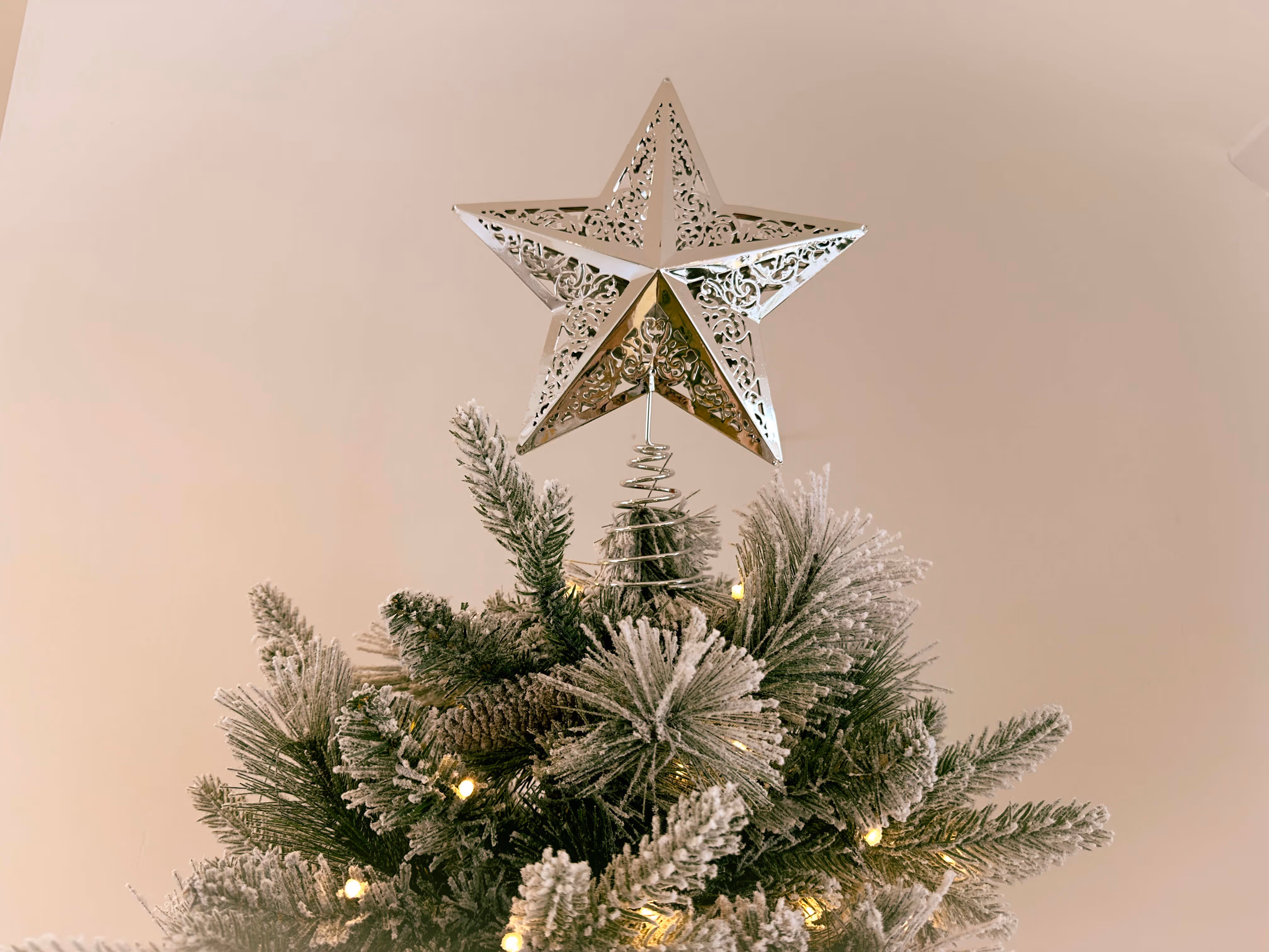  A close-up of a silver star tree topper catching light, placed on top of a fully decorated snow flocked Christmas tree.